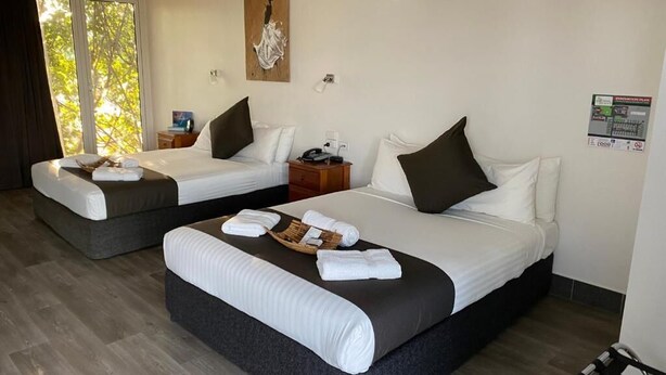 A hotel room with two beds, each with a dark headboard, white bedding, and a small bedside table with a lamp. There is a large window with a view of trees, an air conditioning unit on the wall, and a painting above the window. - Darwin Resort (Winnellie)