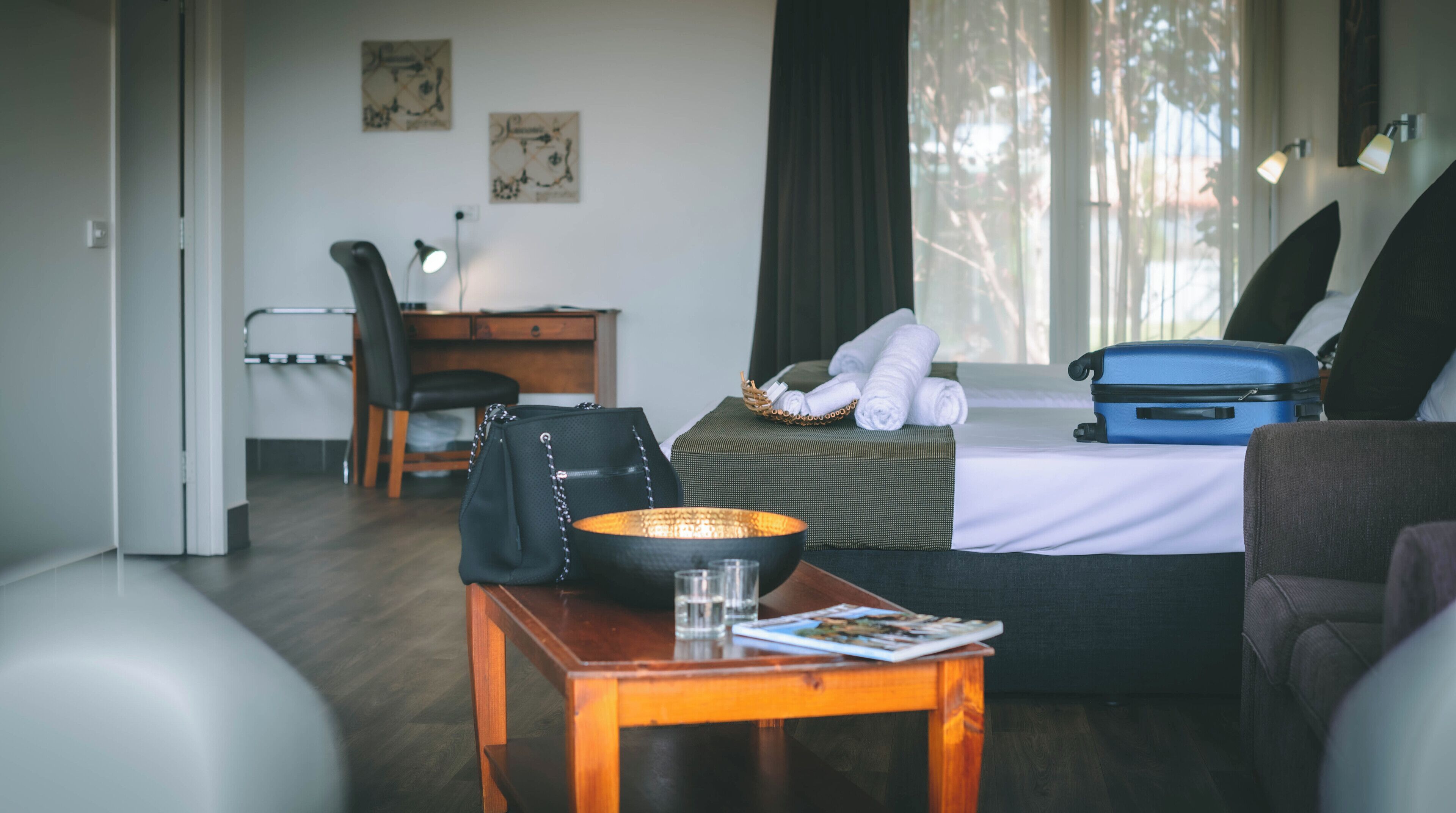Habitación de hotel con cama, escritorio, silla, mesa con un bol y un vaso, y una maleta.