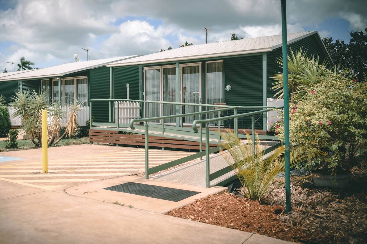 A green building with a ramp leading to a porch, surrounded by plants and a wheelchair-accessible area.
