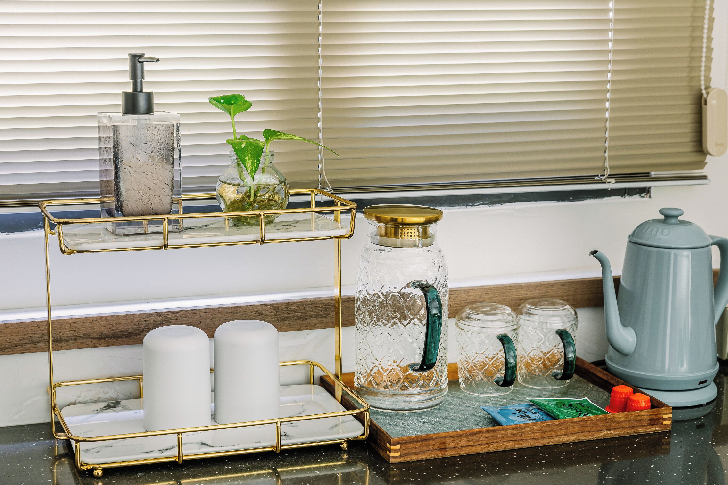 A kitchen counter with a marble tray, a glass dispenser, a kettle, and a plant.