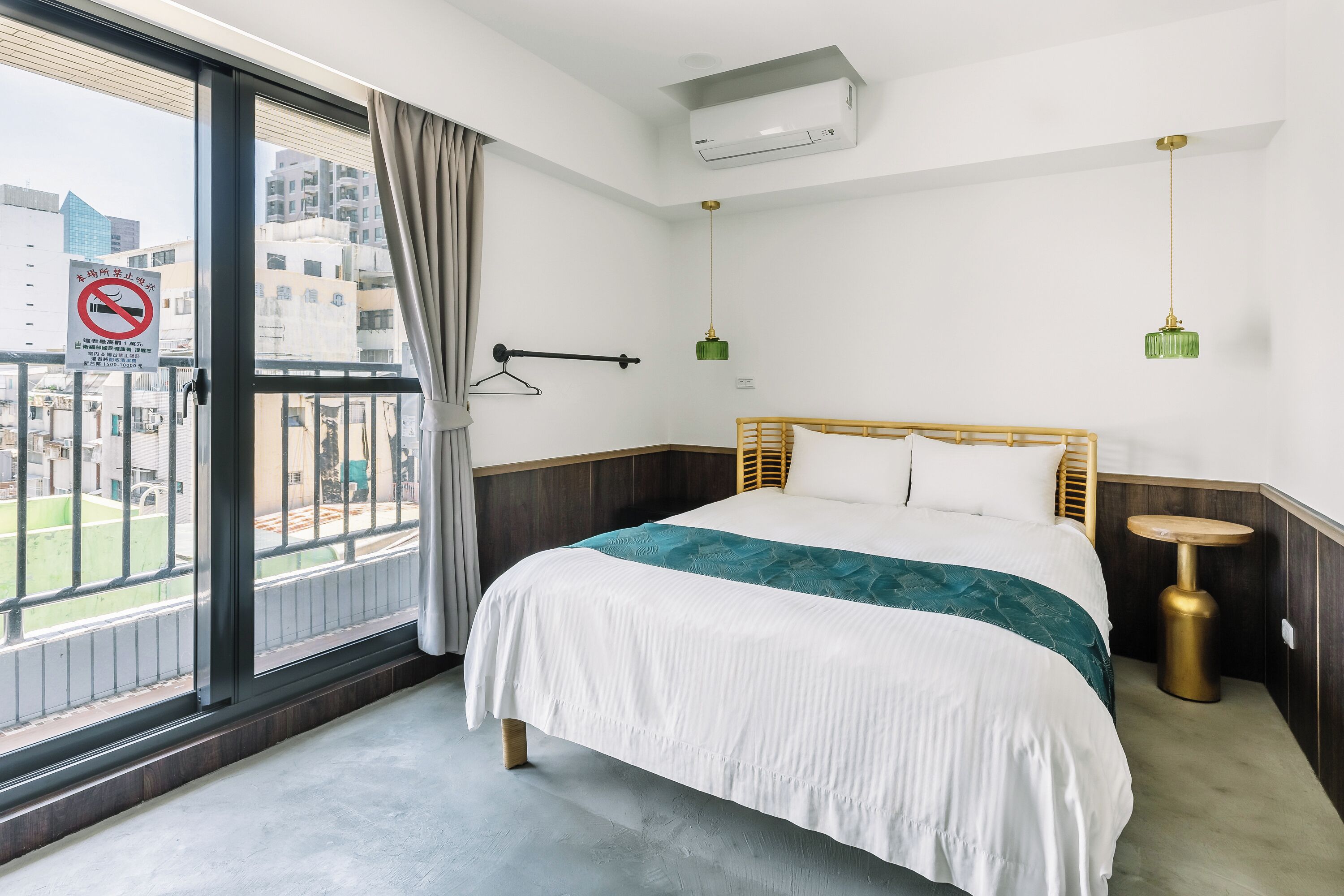 A hotel room with a large window, a bed with a white and teal bedspread, a wooden headboard, a small round table, and a hanging green pendant light.