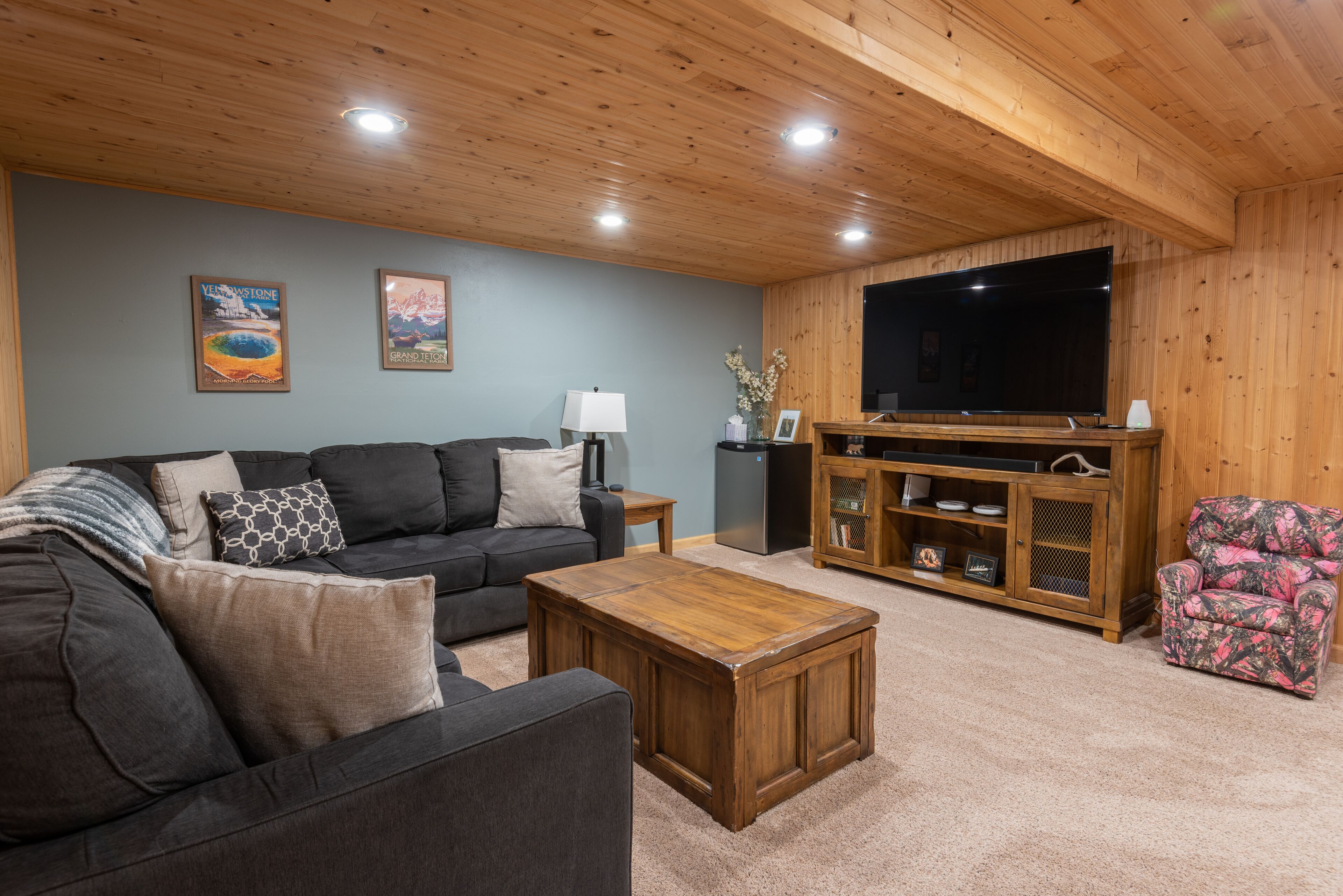 A living room with a dark-colored sofa, a wooden coffee table, a flat-screen TV on a wooden stand, and a patterned armchair.