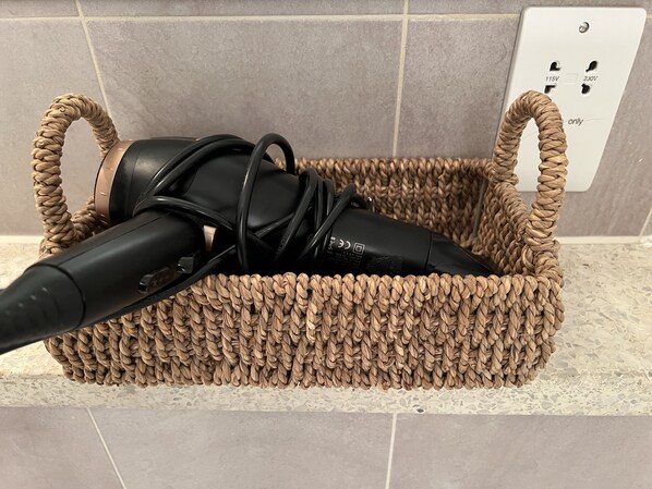 A woven basket containing a hairdryer and a black corded appliance on a tiled floor.