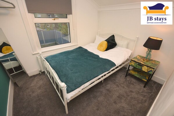 A single-bed room with a white metal frame, a teal blanket, and two pillows. There's a window with a view of a fenced area. A small table with a lamp and a colorful side table with drawers is beside the bed.