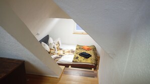 Un pequeño y estrecho dormitorio en el ático con una cama, almohadas, una manta y una mesita con una toalla doblada.