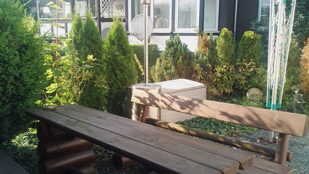 Una mesa de picnic de madera con bancos en un jardín, rodeado de follaje y un edificio con amplias ventanas y una antena parabólica.