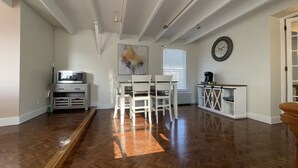 Una habitación amplia con piso de madera, un comedor con sillas blancas, una pequeña cocina con cafetera y un gran cuadro abstracto en la pared.