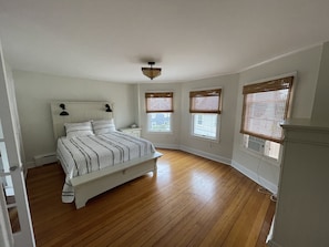 Quarto com uma cama, mesa de cabeceira, ventilador de teto e três janelas com persianas.