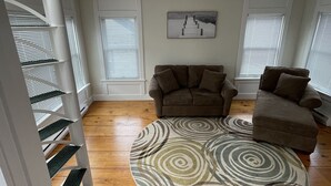 Amplia sala de estar con piso de madera, sofá marrón, alfombra estampada y escalera con barandillas blancas.
