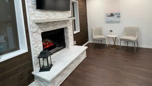 A modern living room with a fireplace, a flat-screen TV mounted above it, and two armchairs facing a small round table with a plant.