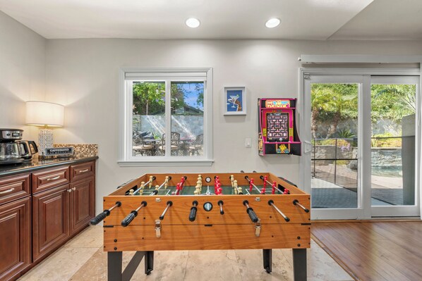 A foosball table in a room with a kitchen area, a dining table with chairs, and a sliding glass door leading to an outdoor area. - Charming Coastal Escape: Resort-Style Walk-In Spa (Oceanside)