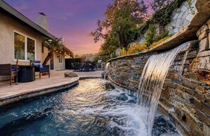A swimming pool with a waterfall feature, outdoor seating, and a stone wall. - Charming Coastal Escape: Resort-Style Walk-In Spa (Oceanside)