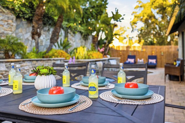 An outdoor dining area with tables set for a meal, surrounded by greenery and a stone wall. - Charming Coastal Escape: Resort-Style Walk-In Spa (Oceanside)