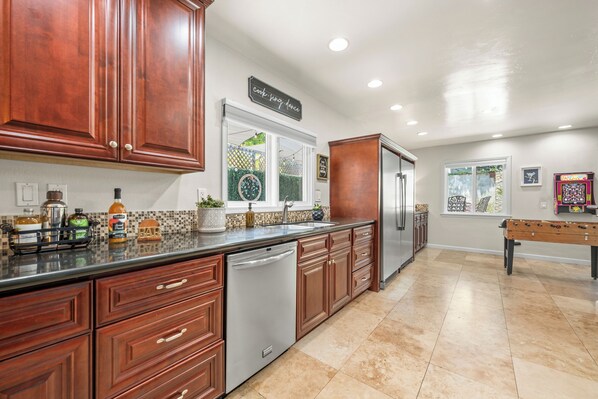 A kitchen with wooden cabinets, a stainless steel dishwasher, and a large window. - Charming Coastal Escape: Resort-Style Walk-In Spa (Oceanside)