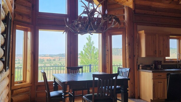 A spacious dining area with a wooden table and chairs, large windows offering a view of the outdoors, and a rustic ceiling with a chandelier. - Large garage for bikes. Beautiful log home.  On golf course. (Hulett)