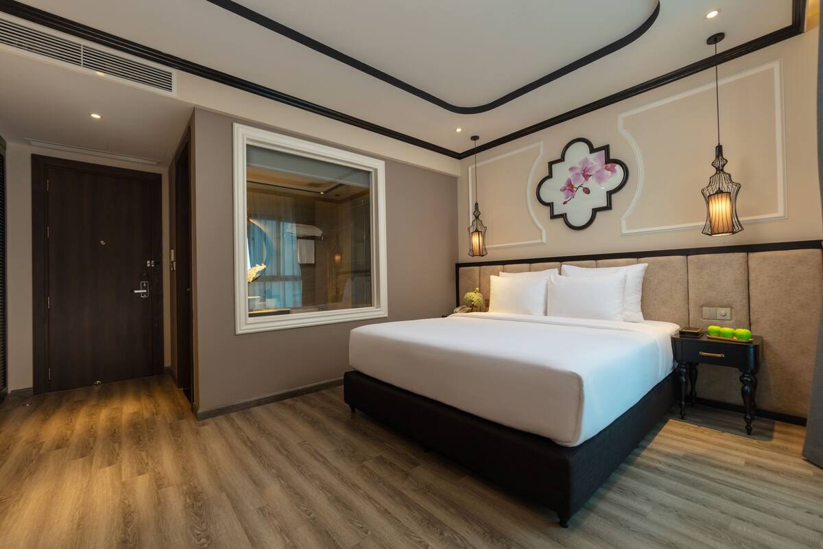 A modern hotel room with a large bed, a window, a nightstand, and a decorative wall piece.