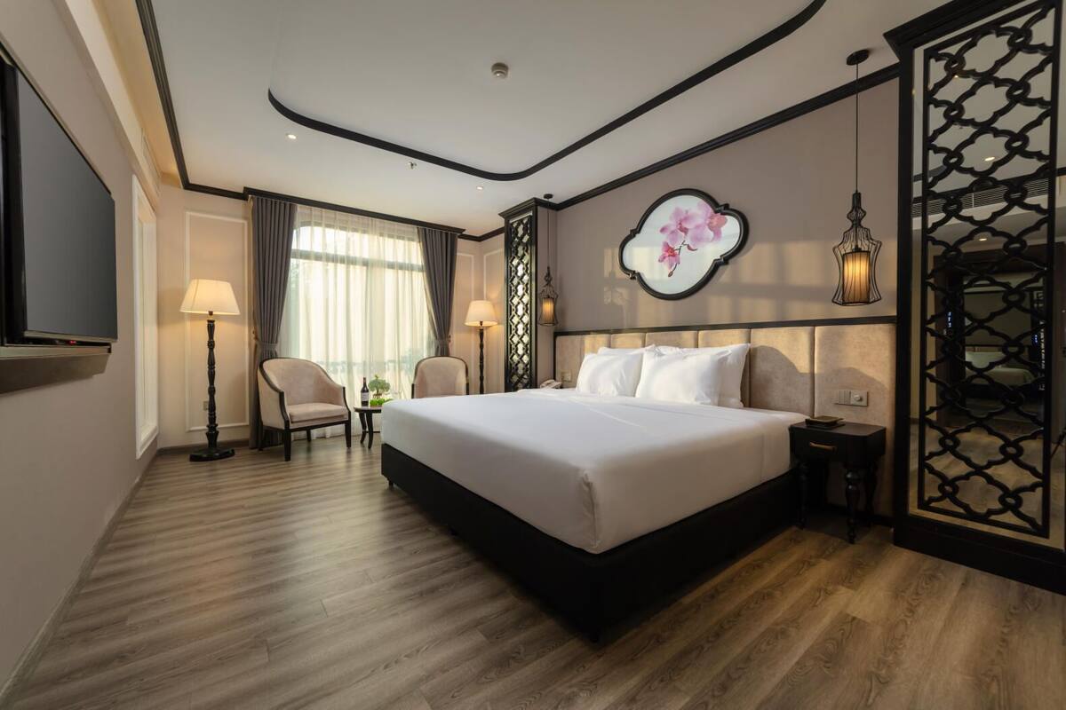 A hotel room with a large bed, two chairs, a TV, and a decorative wall piece.