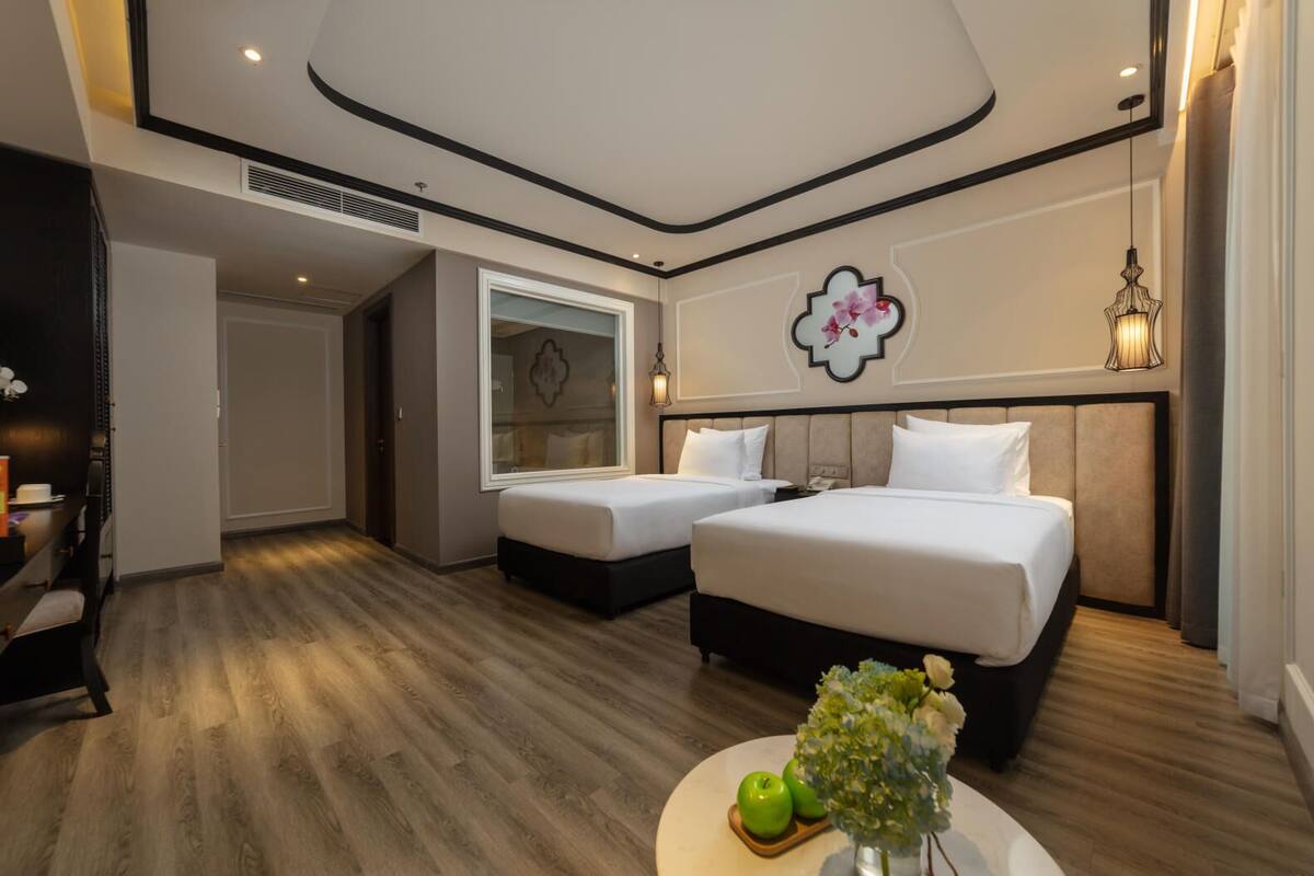 A hotel room with two beds, a round coffee table with apples and flowers, a large window, and a decorative wall piece.