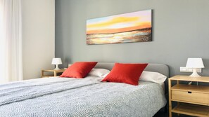 Ein ordentlich bezogenes Bett mit einer gemusterten Decke, zwei roten Kissen und einem Wandbild eines Sonnenuntergangs.