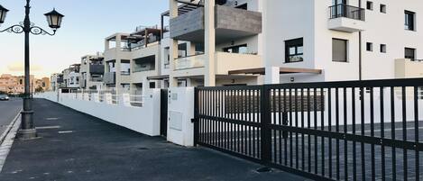 A modern multi-story residential building with balconies, a gated entrance, and a street view.