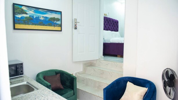 A room with a green armchair, a blue armchair, a bed with a purple headboard, a painting of a landscape, and a microwave. - Charming 1-Bed Apartment in Abuja (Abuja)