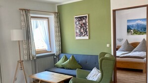 Ein modernes Wohnzimmer mit einem grünen Sofa, einem Holztisch und einem grünen Sessel. Ein Fenster mit Vorhängen, ein gerahmtes Bild an der Wand und ein Bett mit Kissen im angrenzenden Zimmer.