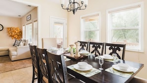 Un comedor con una mesa de madera oscura dispuesta para cuatro personas, rodeada de sillas de madera oscura. Arriba, una araña de candelabros ilumina la mesa, y amplias ventanas dejan entrar luz natural.