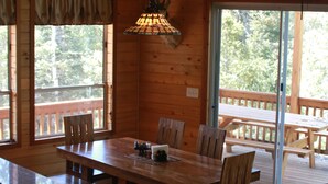 Un interior de madera con una mesa de comedor, sillas, un sofá y un amplio ventanal con vista a árboles.
