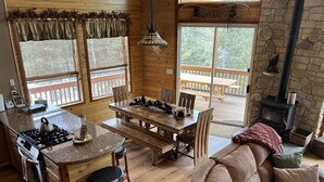 Interior acogedor de una cabaña con zona de comedor de madera, chimenea de piedra y vistas al exterior.