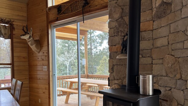 Interior de una cabaña con pared de piedra, muebles de madera, chimenea y vista a una terraza exterior.