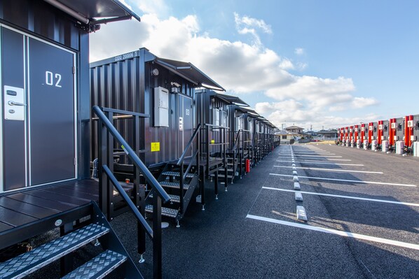 A row of compact, industrial-style cabins with metal railings and numbered doors, arranged in a parking lot with clear demarcations.