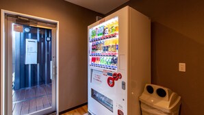 A vending machine with various beverages and snacks, a trash bin, and a door with a mat. - HOTEL R9 The Yard Nakatsu (Nakatsu)