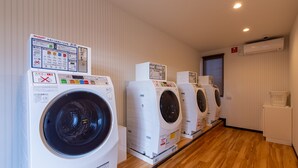 A laundry facility with multiple washing machines lined up against a wall, wooden flooring, and an air conditioning unit on the wall. - HOTEL R9 The Yard Nakatsu (Nakatsu)