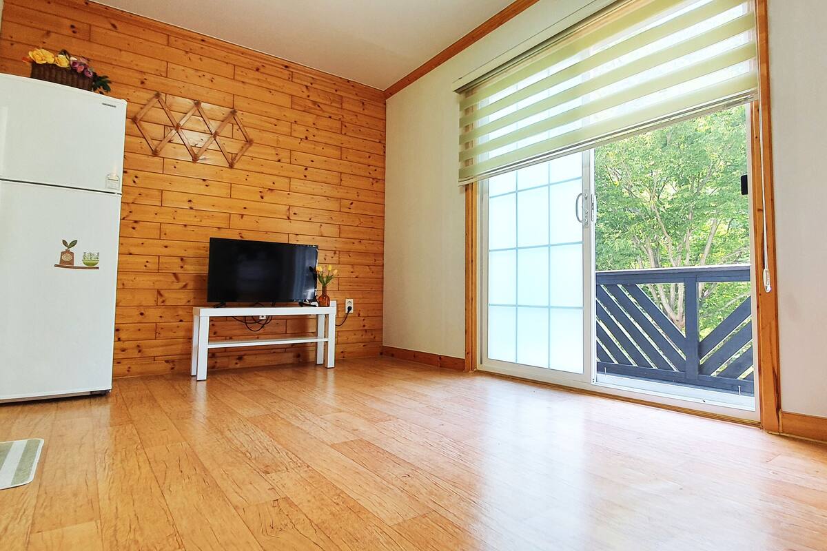 Een kamer met houten wanden, een witte koelkast, een televisie op een standaard, een grote schuifglazen deur naar een balkon en een stapel opgevouwen dekens.