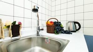 Una cocina con un lavamanos de acero inoxidable, un grifo, una tetera, una cesta de frutas y un florero con flores.