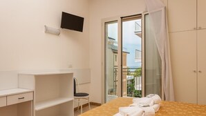 Een hotelkamer met een bed, een bureau, een stoel, een televisie en een balkon met uitzicht op gebouwen en de zee.