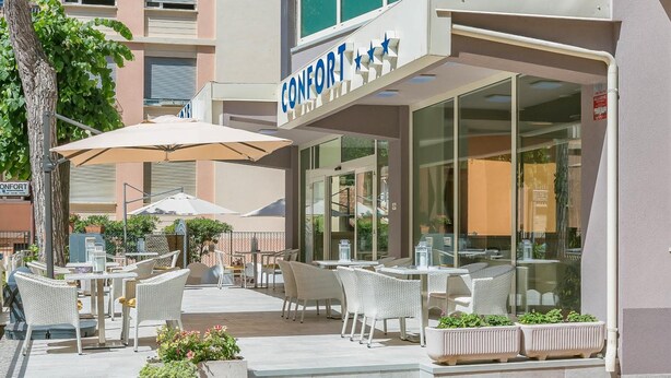 Een terras met rieten meubels en parasols, een gebouw met een 'CONFORT'-bord en een heldere lucht.