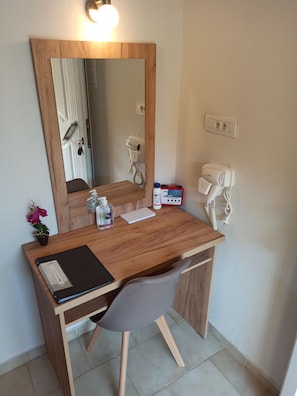 A compact wooden desk with a mirror, a hairdryer, and a bottle of hand sanitizer.