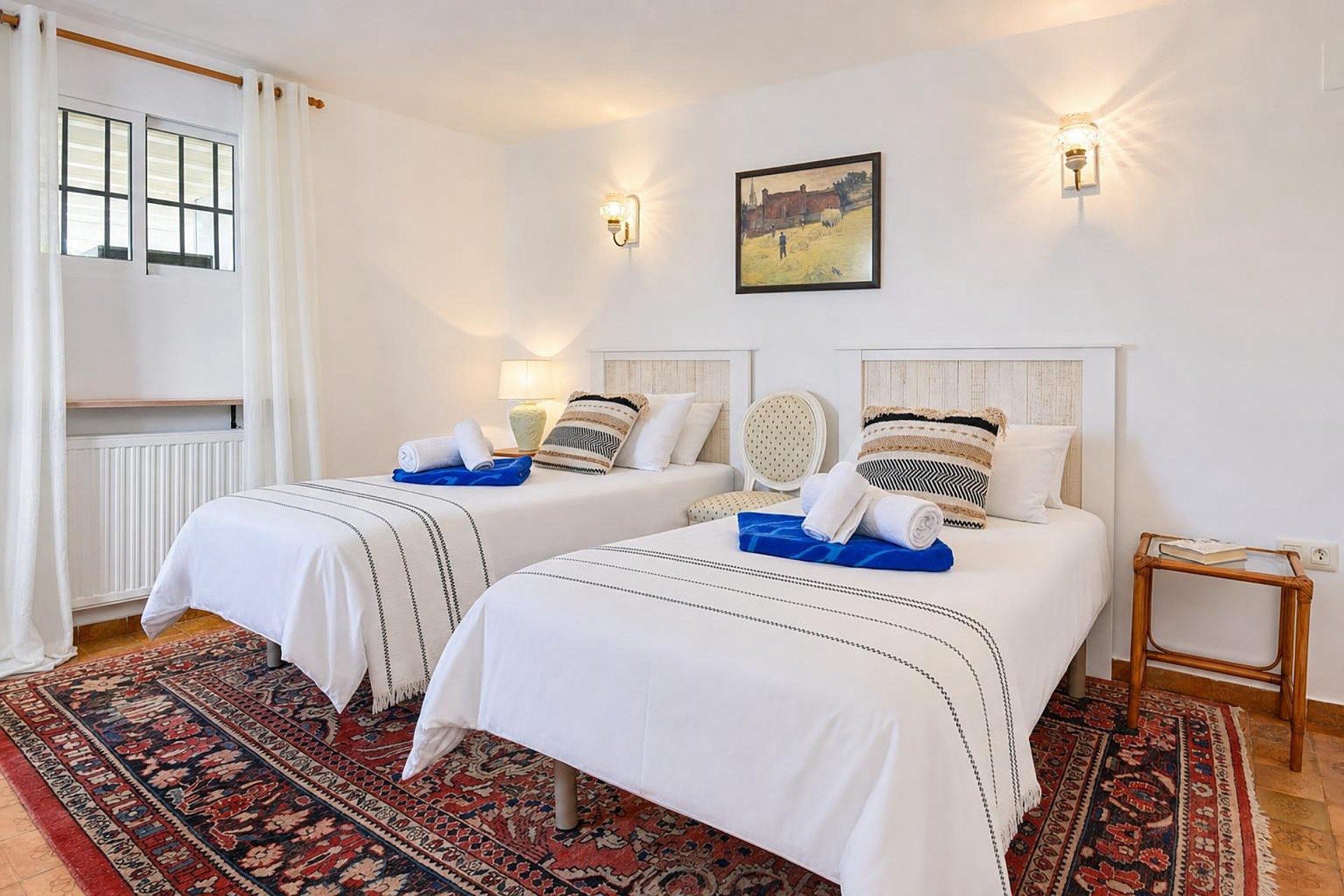 A hotel room with two beds, a patterned rug, a painting on the wall, and a small wooden side table.