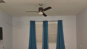 Una habitación con ventilador de techo, ventana con persianas y piso alfombrado.