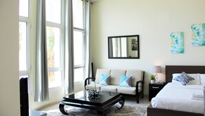 A hotel room with a sofa, a bed, a nightstand with a lamp, a mirror, and artwork on the wall. - IRN - Spacious Studio in Discover Garden (Dubai)