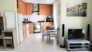 A compact kitchen with wooden cabinets, a refrigerator, a stove, and a microwave. There is a dining table with chairs and a flat-screen TV on a stand. - IRN - Spacious Studio in Discover Garden (Dubai)