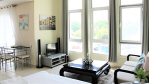 A hotel room with a bed, a TV, a dining table with chairs, and large windows with curtains. - IRN - Spacious Studio in Discover Garden (Dubai)