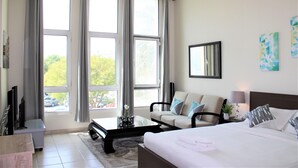A hotel room with a bed, a sofa, a coffee table, a TV, and large windows. - IRN - Spacious Studio in Discover Garden (Dubai)