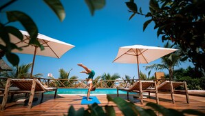 Zona de piscina con sillones reclinables de madera, dos sombrillas grandes y una persona haciendo una postura de yoga sobre una colchoneta.