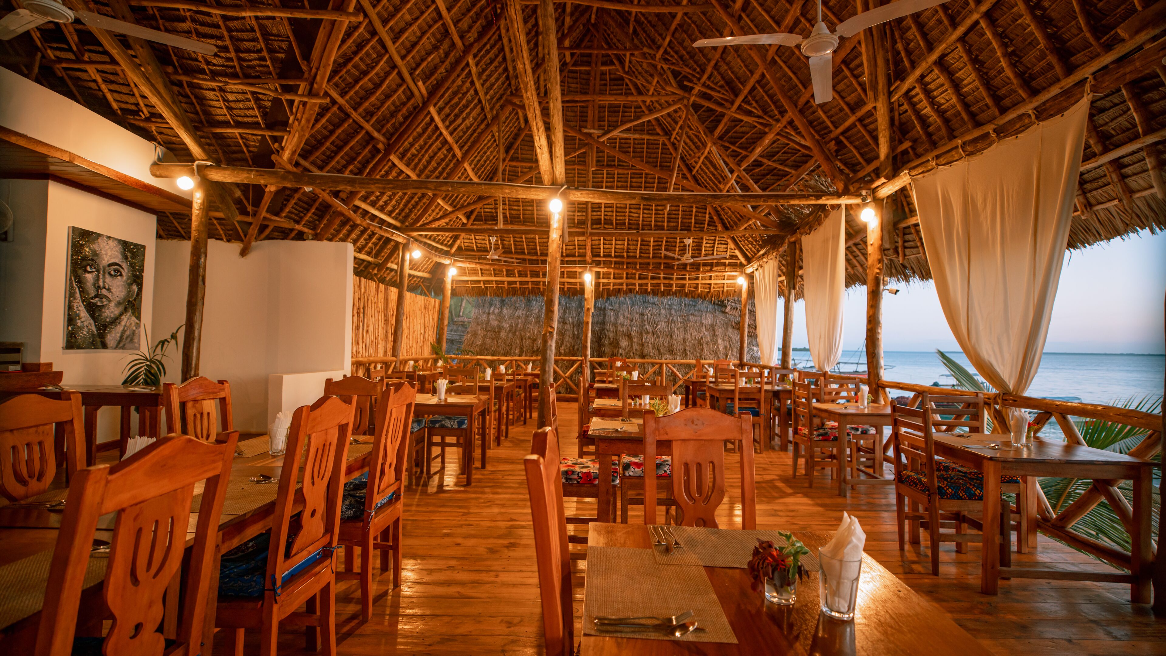 A restaurant with wooden furniture and a thatched roof, overlooking the sea.