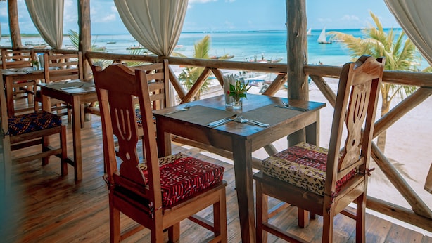 Un área de comedor al aire libre con mesas y sillas de madera, techo de paja y vista al océano.