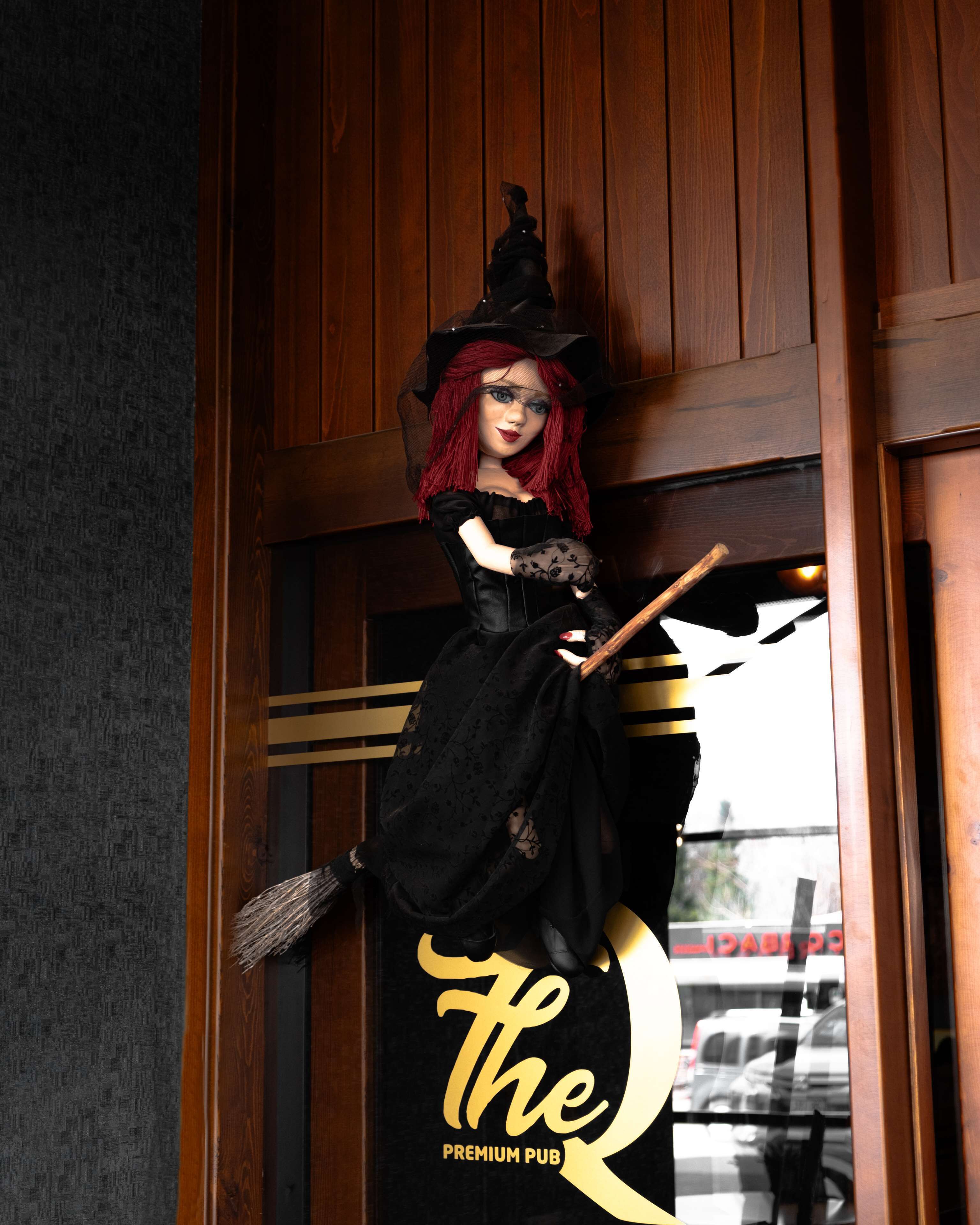 A doll with red hair and a black dress, positioned in front of a door with a sign reading "The Q Premium Pub".