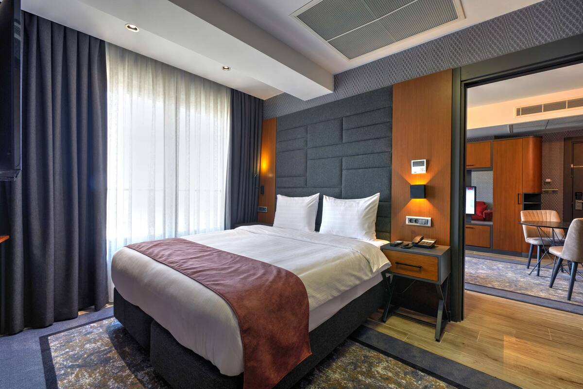 Een moderne hotelkamer met een groot bed, een bureau met lamp en uitzicht op de aangrenzende eetruimte.