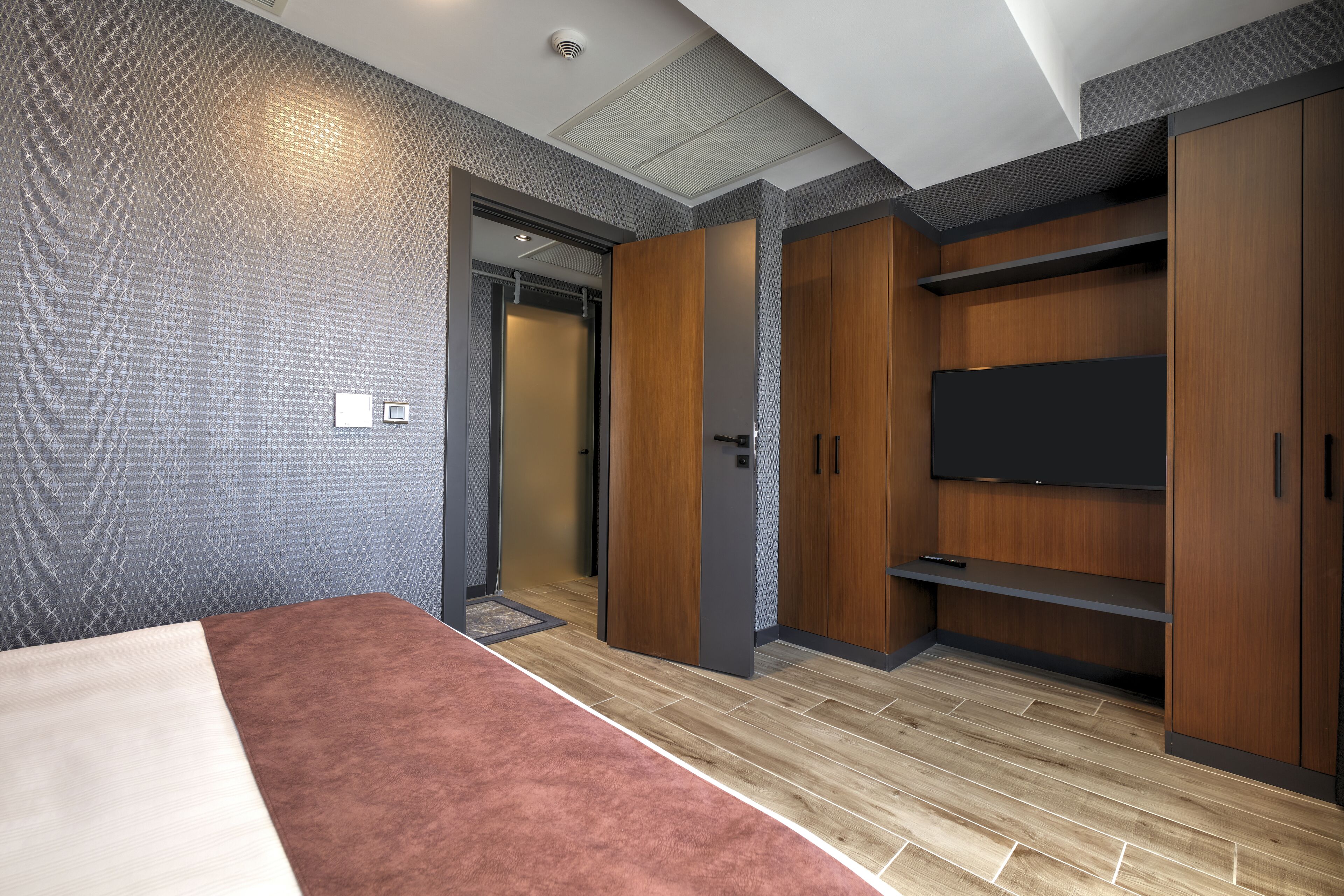 A modern hotel room with a large bed, wooden wardrobe, built-in TV, and a patterned wall.